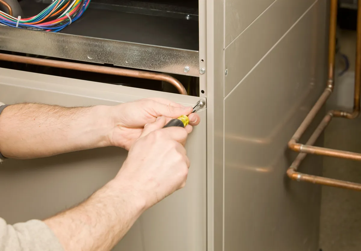 A technician using a screwdriver to tighten a screw on the side of a furnace, with visible copper pipes.