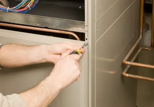 A technician using a screwdriver to tighten a screw on the side of a furnace, with visible copper pipes.