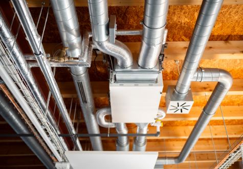 Exposed commercial HVAC ductwork and ventilation unit mounted to a ceiling with visible metal pipes, beams, and utility lines.