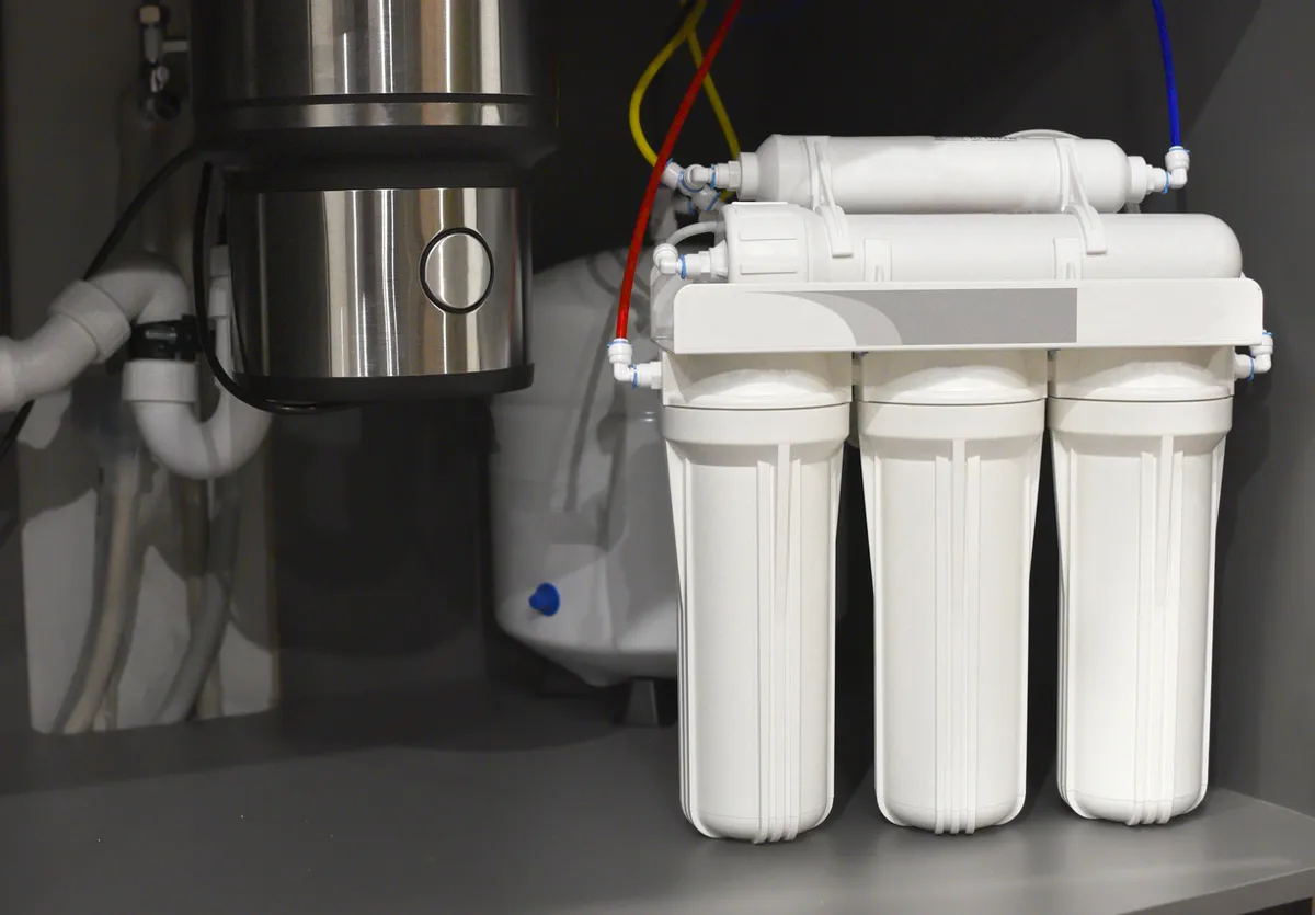 Under-sink water filtration system with multiple white filter canisters connected by colored tubing, installed beside a garbage disposal unit and plumbing pipes inside a kitchen cabinet.