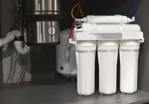 Under-sink water filtration system with multiple white filter canisters connected by colored tubing, installed beside a garbage disposal unit and plumbing pipes inside a kitchen cabinet.