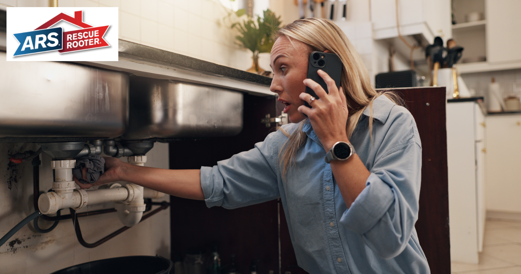 A woman kneeling in her kitchen, calling ARS to request a plumbing service call while holding a rag to control a water leak under the sink. 