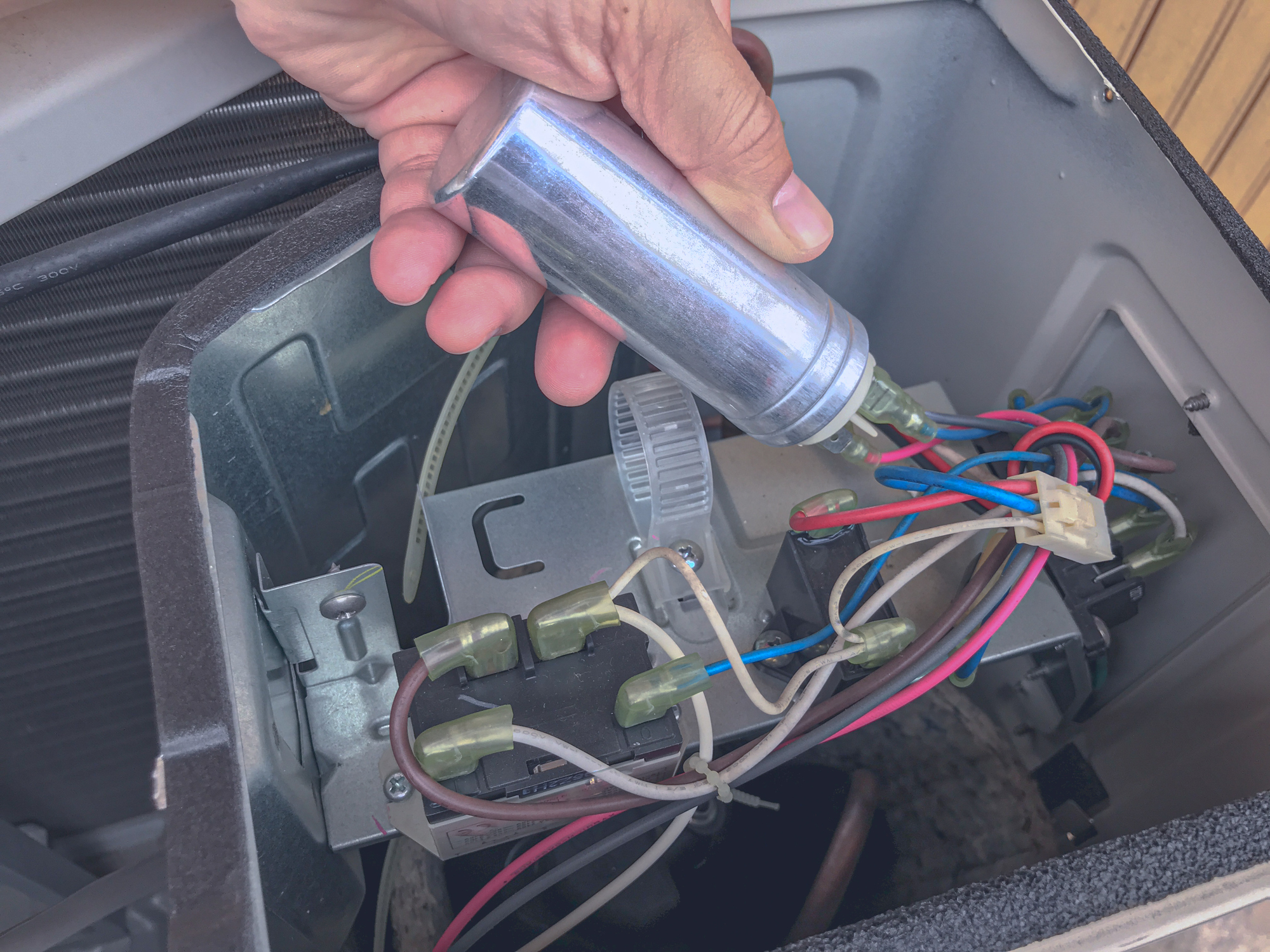 ARS technician checking an outdoor AC system for signs of a bad AC capacitor part, holding it above the system wiring components. 